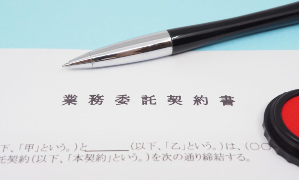 業務委託契約書の書類とペン、印鑑が並べられている。契約内容による残業代支払い回避の事例を示唆。