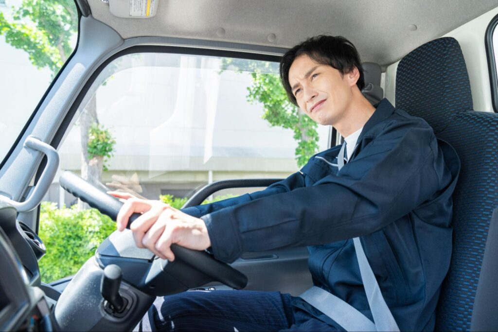 トラックを運転する若い男性ドライバー。やや疲れた表情でハンドルを握っている様子。