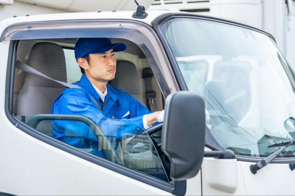 トラックを運転する男性ドライバー。青い作業服とキャップを着用して真剣な表情で運転中。