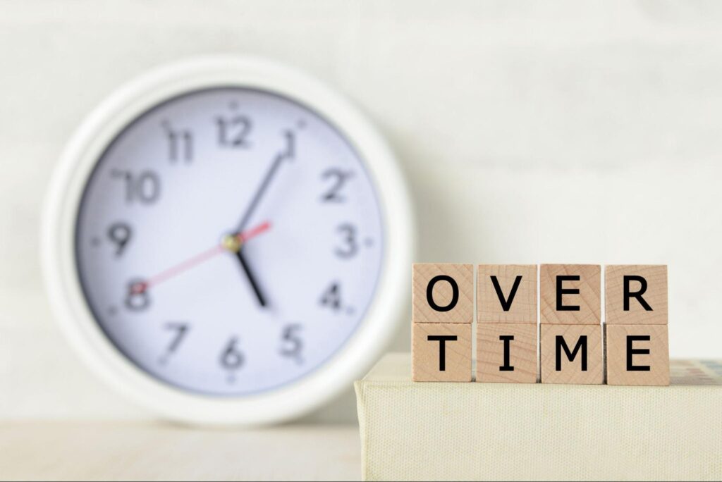 時計と“OVERTIME”と書かれた木製ブロック。残業時間の概念を視覚的に示したイメージ。